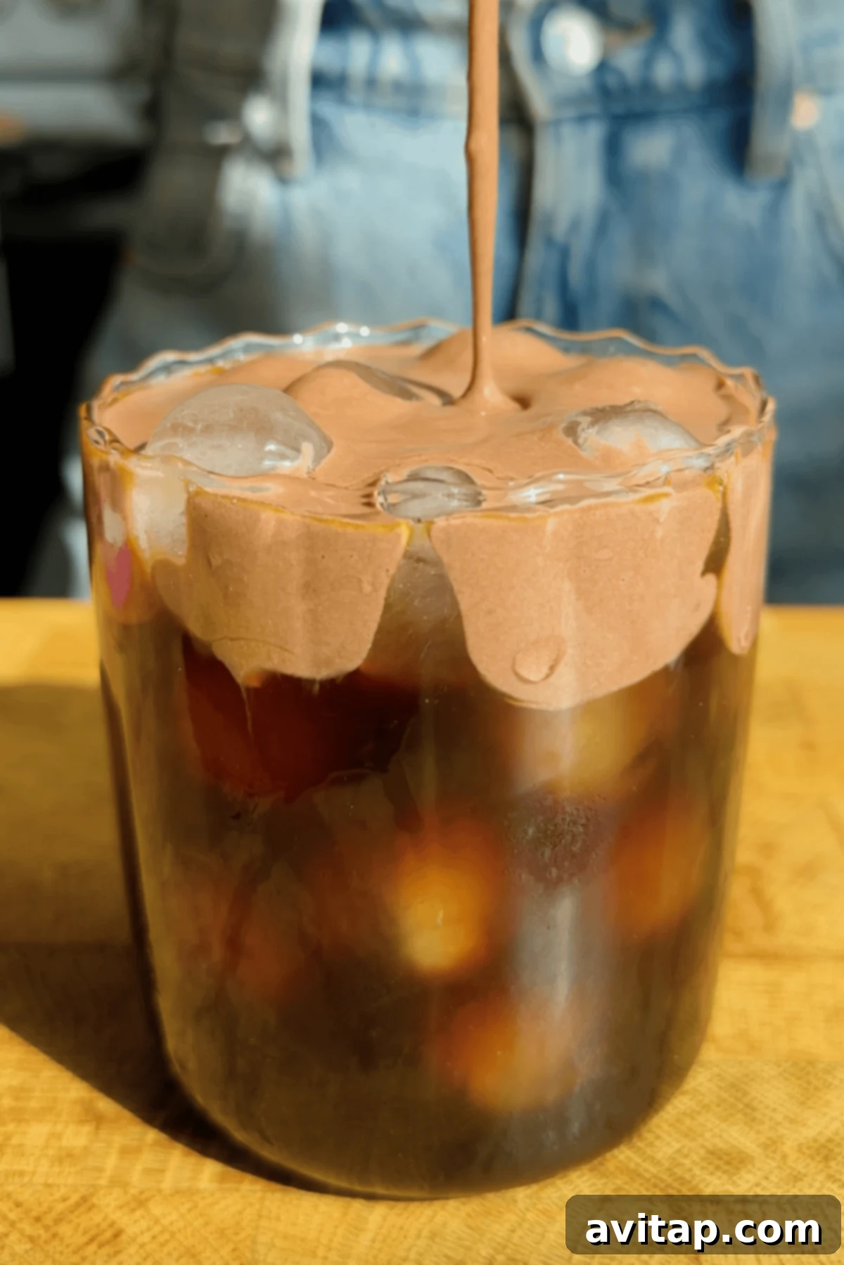 Starbucks Protein-Powered Cold Foam 2 A tall glass of iced coffee, crowned with a generous swirl of chocolate protein cold foam. A hand is gently pouring more of the luscious foam onto the drink, with a soft denim background visible, creating an inviting and delicious scene.