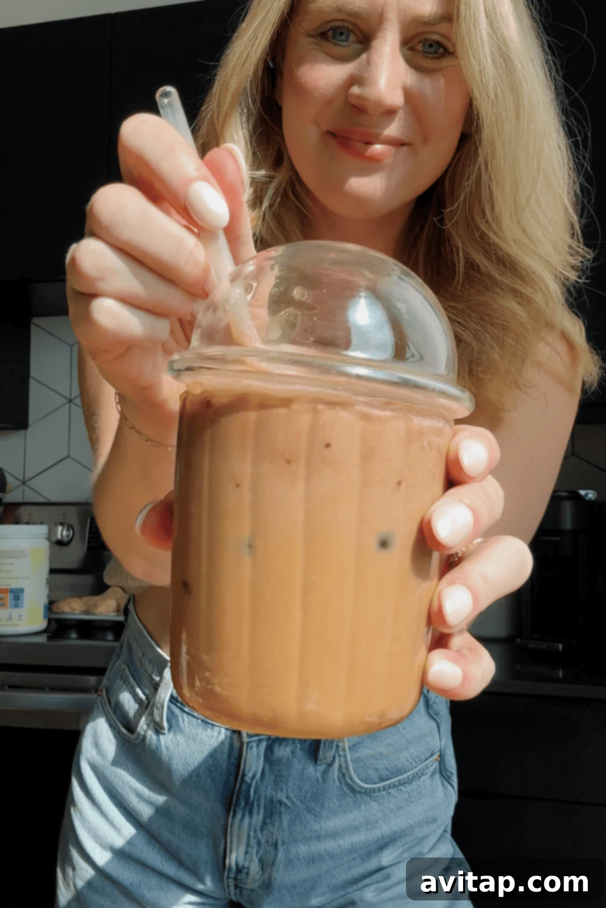 Starbucks Protein-Powered Cold Foam 3 A woman holding a clear plastic cup filled with iced coffee, topped with a generous dome of protein cold foam and a straw, in a brightly lit kitchen. The image conveys refreshment and homemade quality.