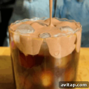 A close-up of iced coffee being topped with a thick, whipped chocolate protein cold foam, highlighting the creamy texture and rich color. A blurred denim background adds a casual touch.