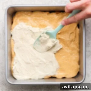 A hand uses a blue spatula to spread a white creamy layer over a yellow batter in a square metal baking pan.