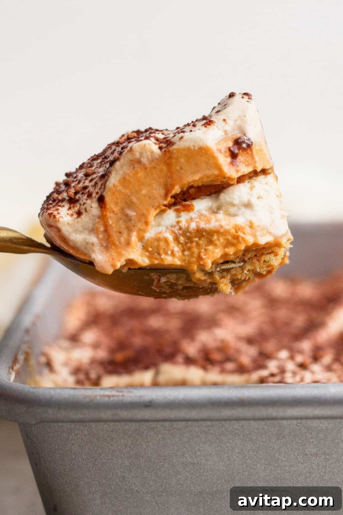 A spoon lifts a layered dessert with a creamy top dusted with cocoa powder from a baking pan.
