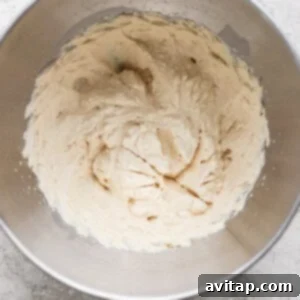 A metal mixing bowl containing light-colored creamed mixture, likely butter and sugar, on a textured gray surface.