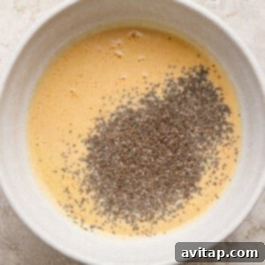 A white bowl containing whisked eggs with a pile of chia seeds on top, set on a light-colored textured surface.