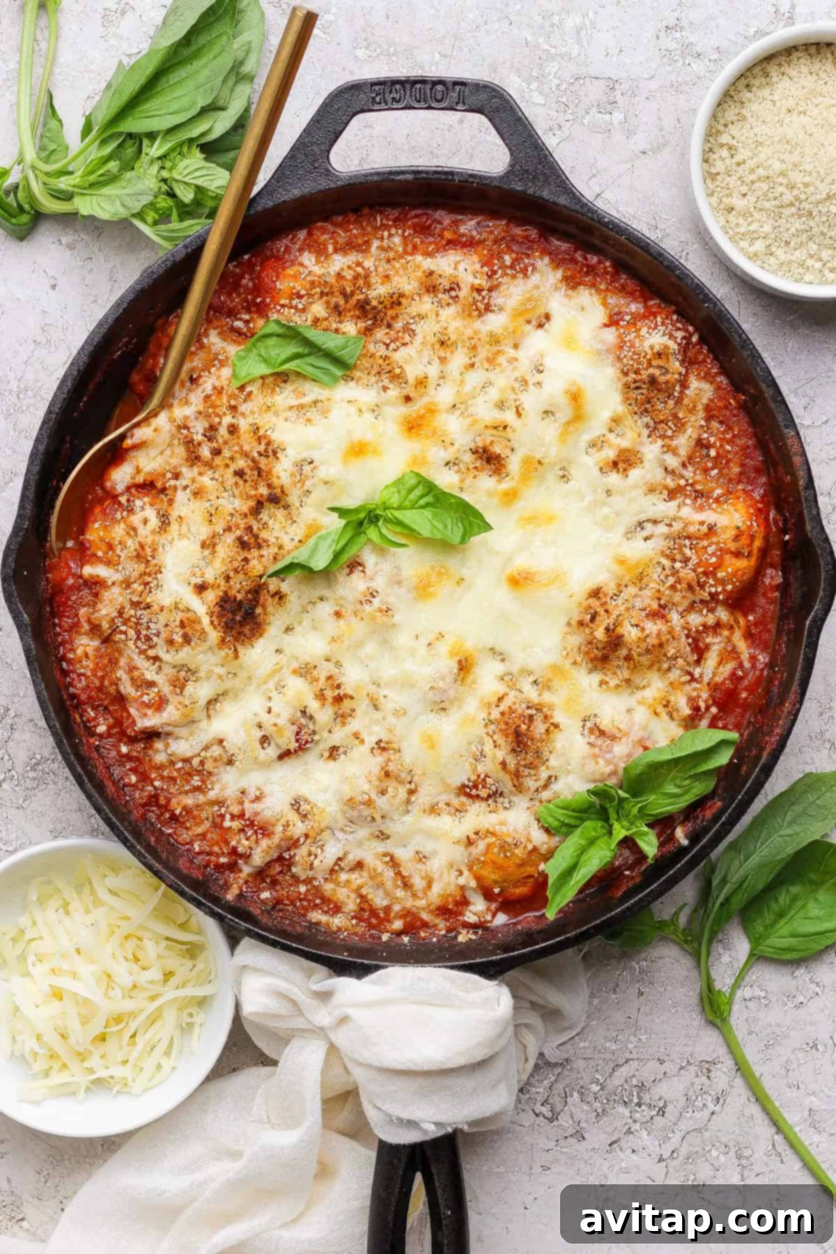 A cast iron skillet filled with baked cheesy tomato dish, garnished with fresh basil, surrounded by small bowls of shredded cheese and breadcrumbs.