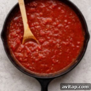 A cast iron skillet filled with chunky tomato sauce, with a wooden spoon resting inside, on a light textured surface.