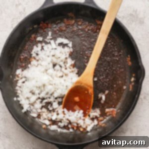 Chopped onions being sautéed in a cast iron skillet with a wooden spoon; residue from previous cooking visible in pan.