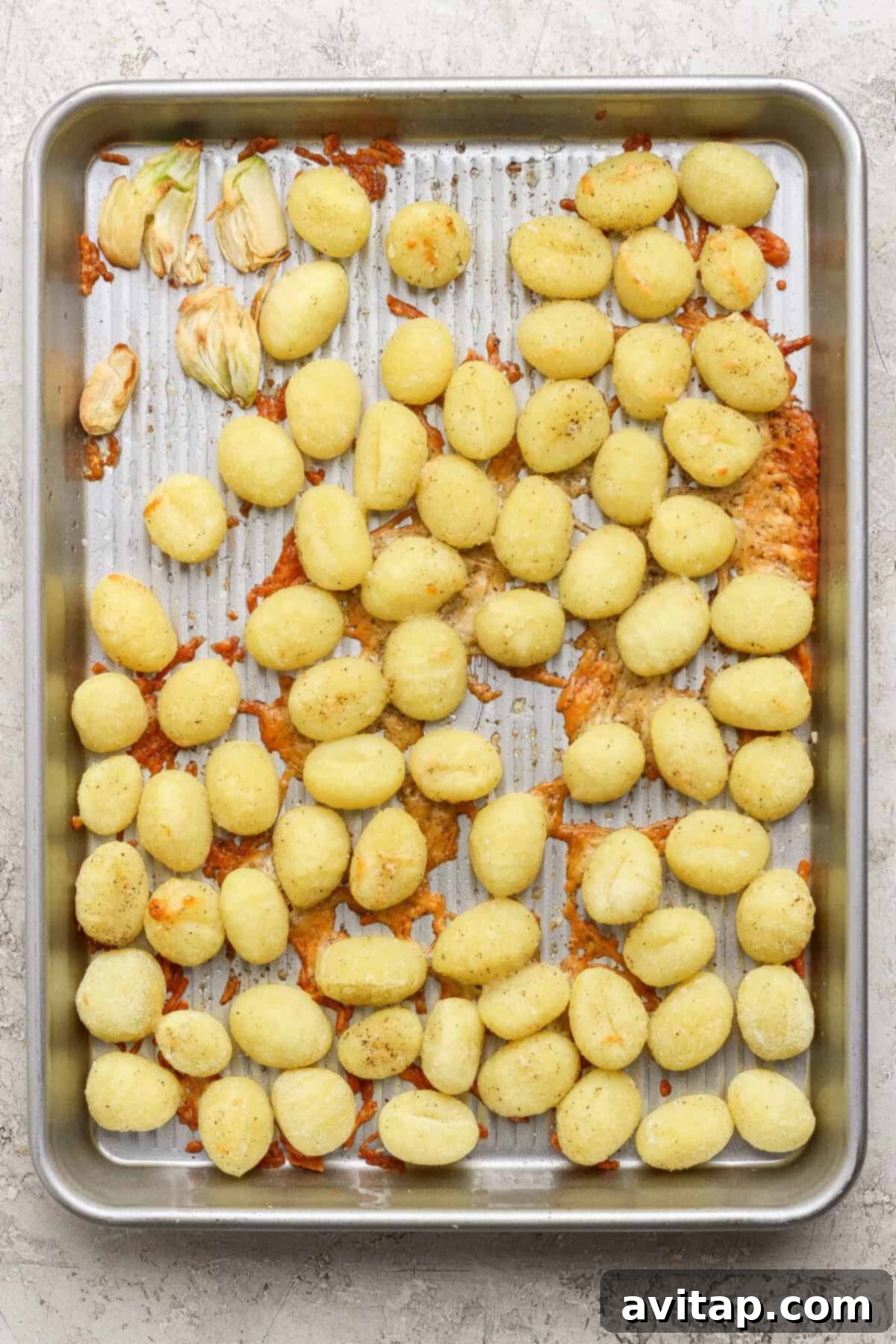 A metal baking sheet with scattered cooked gnocchi, some clumps of melted cheese, and a few roasted garlic cloves on a textured surface.