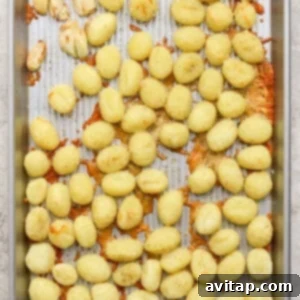 A metal baking sheet with scattered cooked gnocchi, some clumps of melted cheese, and a few roasted garlic cloves on a textured surface.