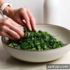 A person massages chopped kale in a large bowl, with a white bottle in the background on a light-colored surface.