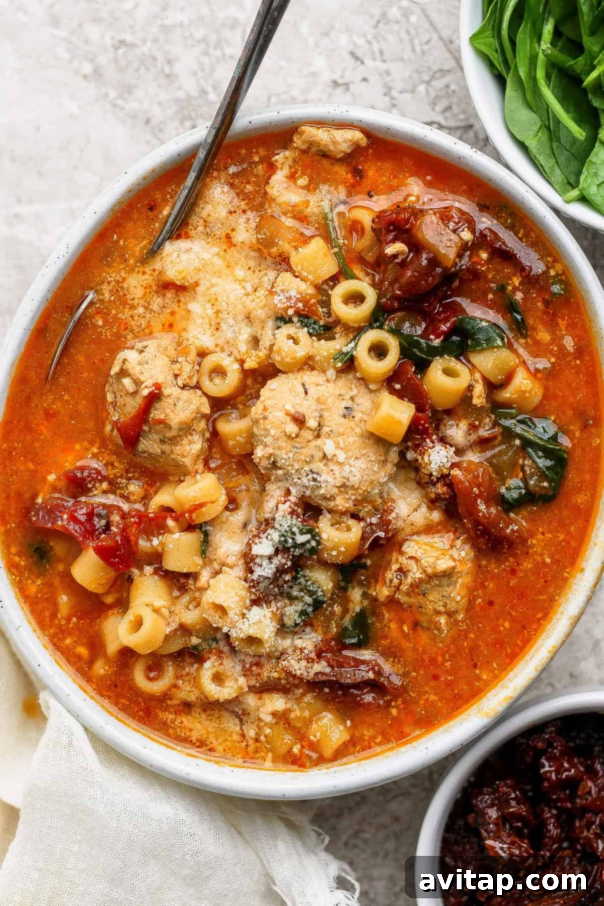 A bowl of tomato-based soup with pasta, chicken, spinach, sun-dried tomatoes, and grated cheese, with a spoon resting inside and fresh spinach on the side.
