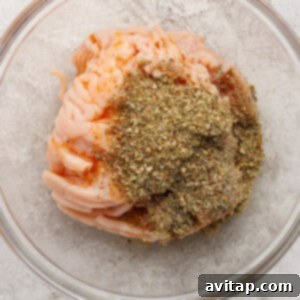 Raw ground meat topped with dried herbs and spices in a clear glass bowl on a light textured surface.