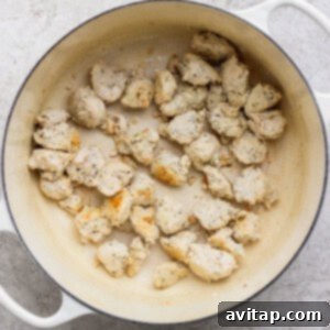 Chunks of cooked chicken are scattered in a white enameled pot on a light textured surface.