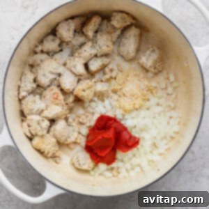 A white pot contains cooked chunks of sausage, chopped onions, minced garlic, and tomato paste on a light countertop.
