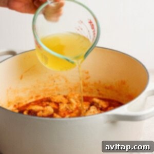 A hand pours chicken broth from a glass measuring cup into a white pot filled with cooked meat and sauce.
