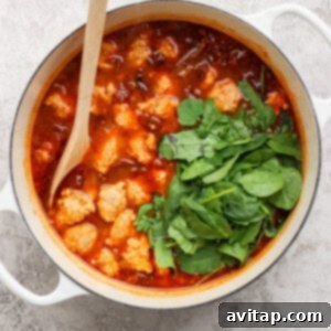 A white pot filled with tomato-based stew, meatballs, and fresh spinach, stirred with a wooden spoon on a light textured surface.