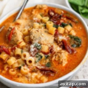 A bowl of soup with meatballs, ditalini pasta, spinach, sun-dried tomatoes, and grated cheese, served with a spoon.