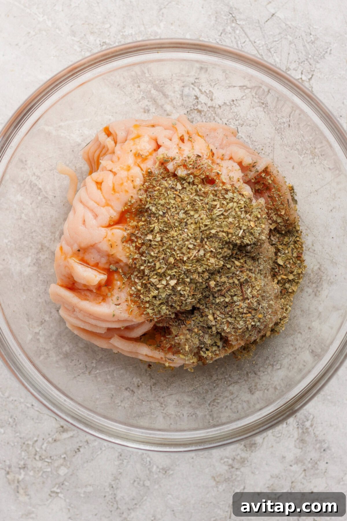 Raw ground meat topped with dried herbs and spices in a clear glass bowl on a light textured surface.