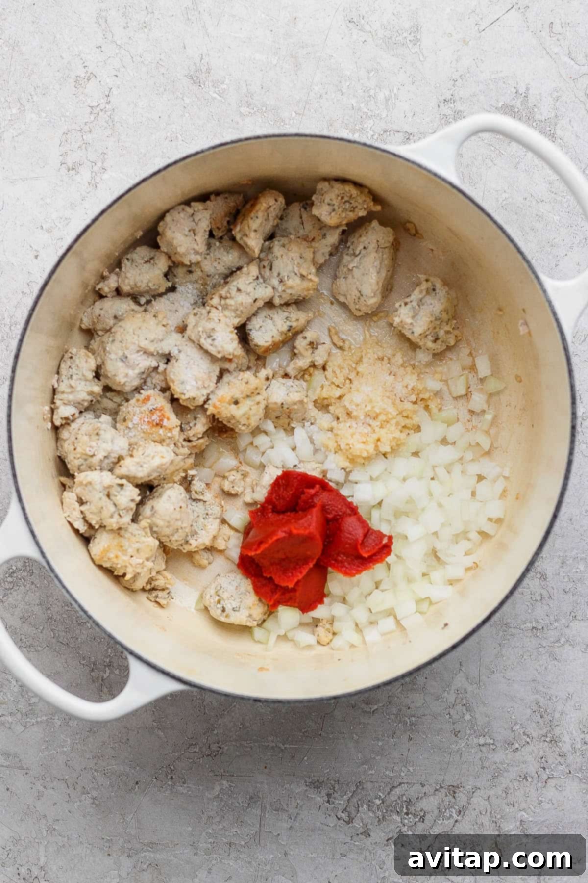 A white pot contains cooked chunks of sausage, chopped onions, minced garlic, and tomato paste on a light countertop.