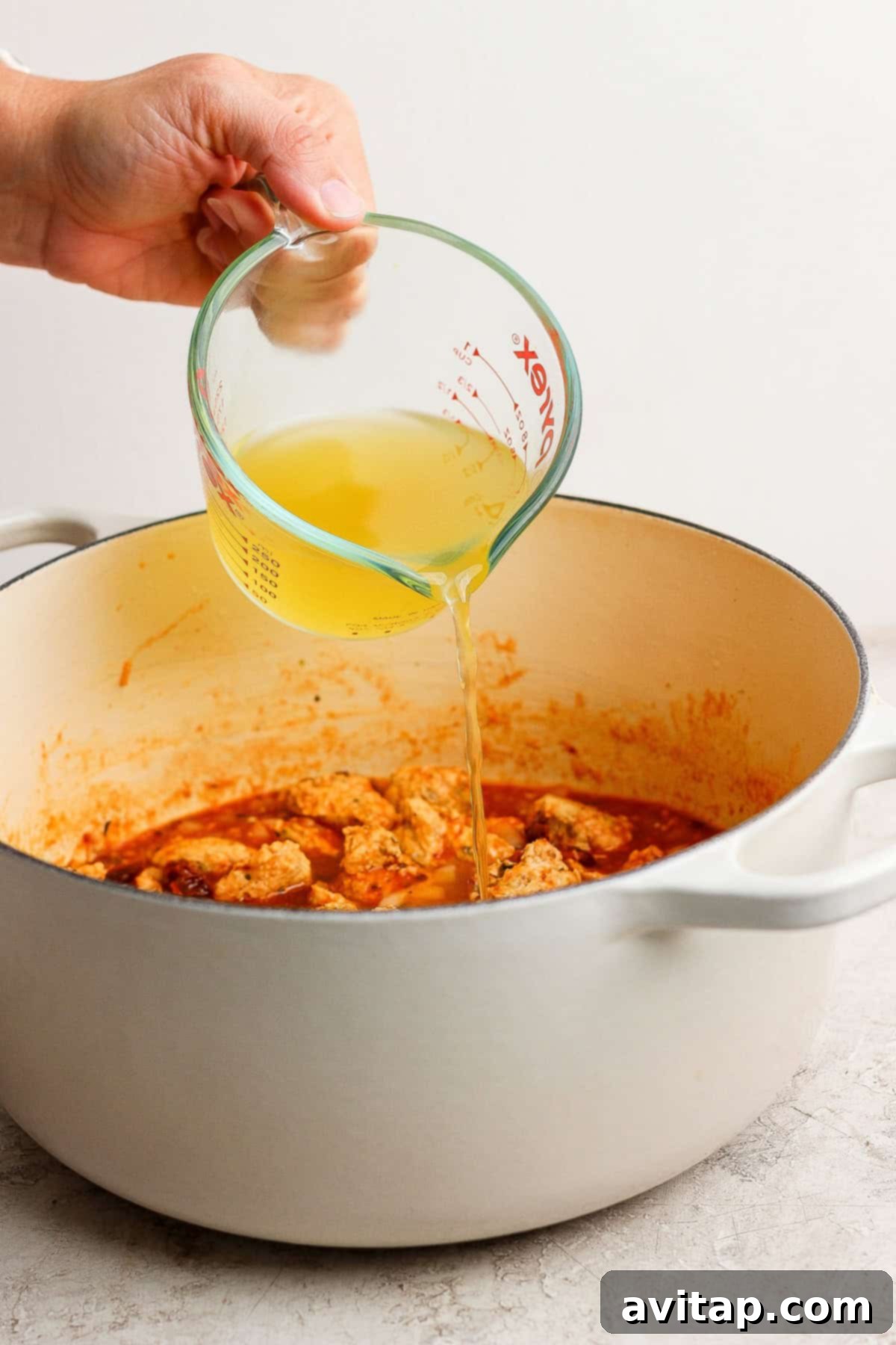 A hand pours chicken broth from a glass measuring cup into a white pot filled with cooked meat and sauce.