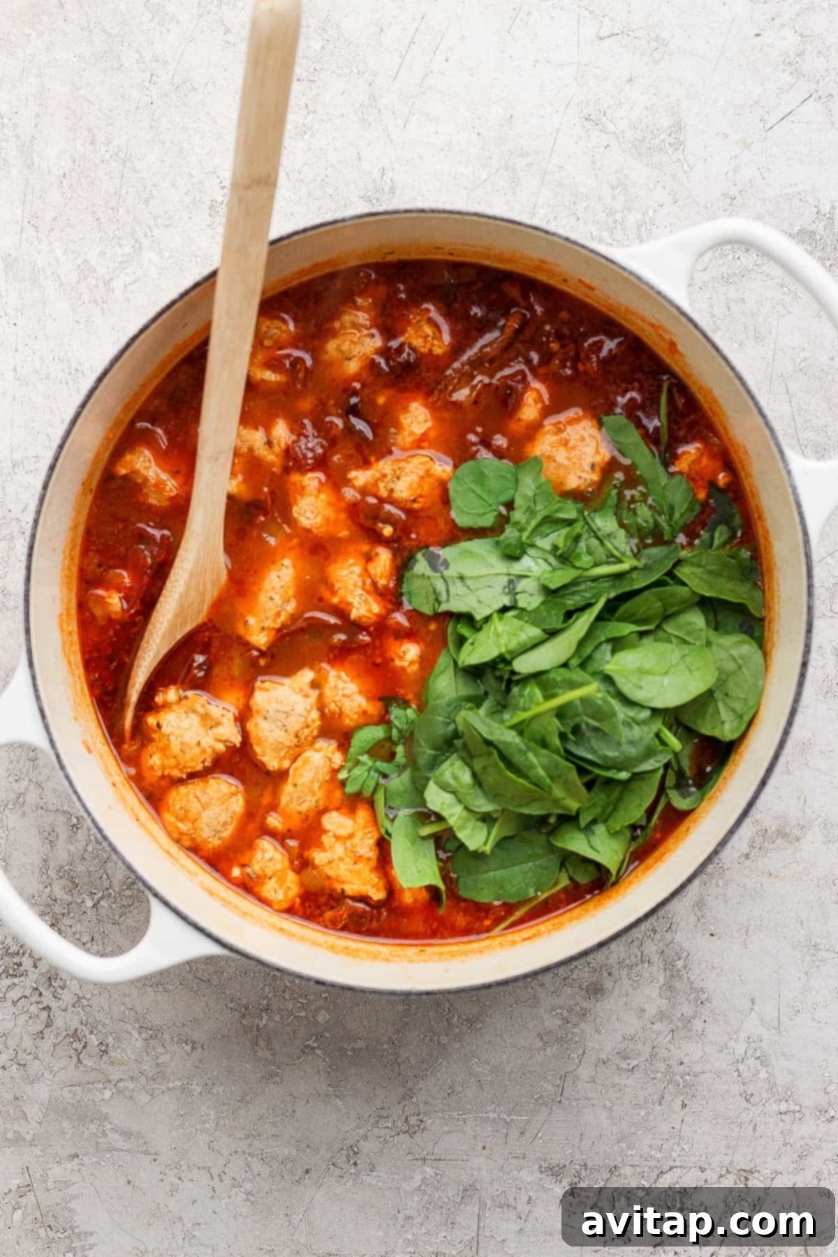 A white pot filled with tomato-based stew, meatballs, and fresh spinach, stirred with a wooden spoon on a light textured surface.
