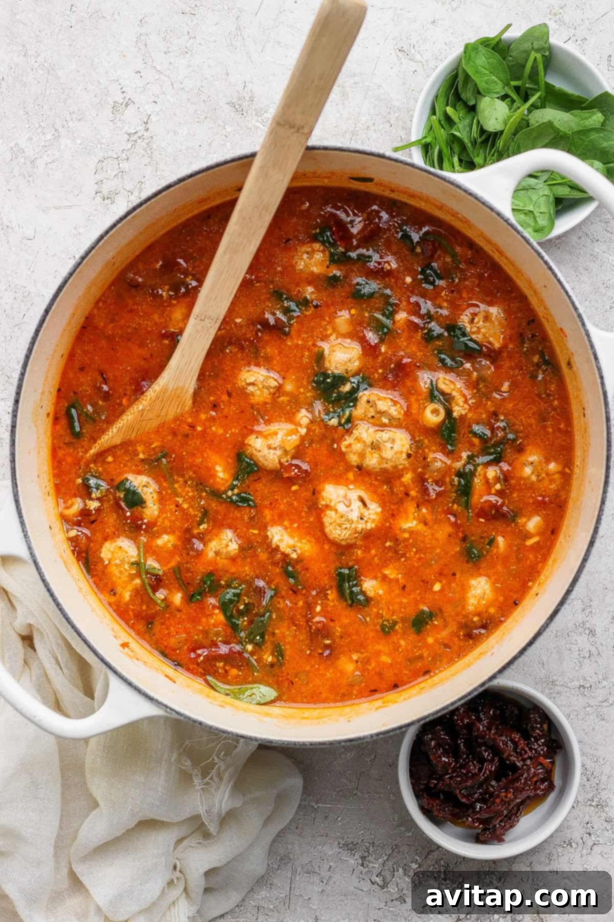A pot of tomato-based soup with meatballs and spinach, a wooden spoon resting inside. Nearby are bowls of fresh spinach and sun-dried tomatoes.