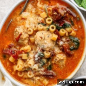 A bowl of tomato-based soup with meatballs, ditalini pasta, sun-dried tomatoes, spinach, and grated cheese on top.