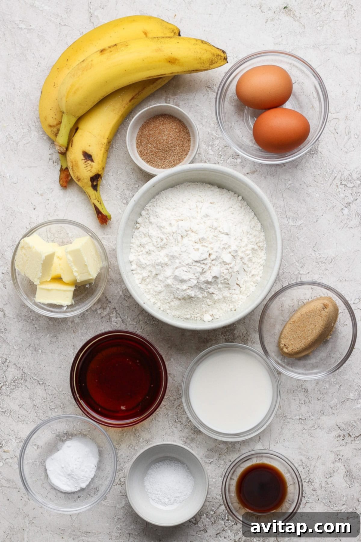 Essential ingredients for a delicious banana bread recipe: ripe bananas, farm-fresh eggs, all-purpose flour, granulated sugar, unsalted butter, milk, brown sugar, baking powder, pure vanilla extract, a pinch of salt, and warm cinnamon, all artfully arranged in individual bowls on a bright, inviting surface.