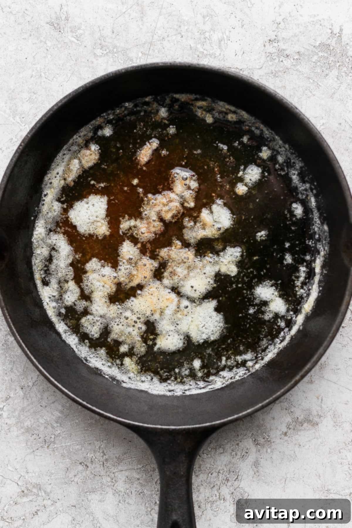 A cast iron skillet holds a pool of beautifully browned butter with a delicate layer of foam, positioned on a light, textured surface. This essential ingredient is perfectly prepared and ready to be incorporated into your next delicious banana bread recipe.