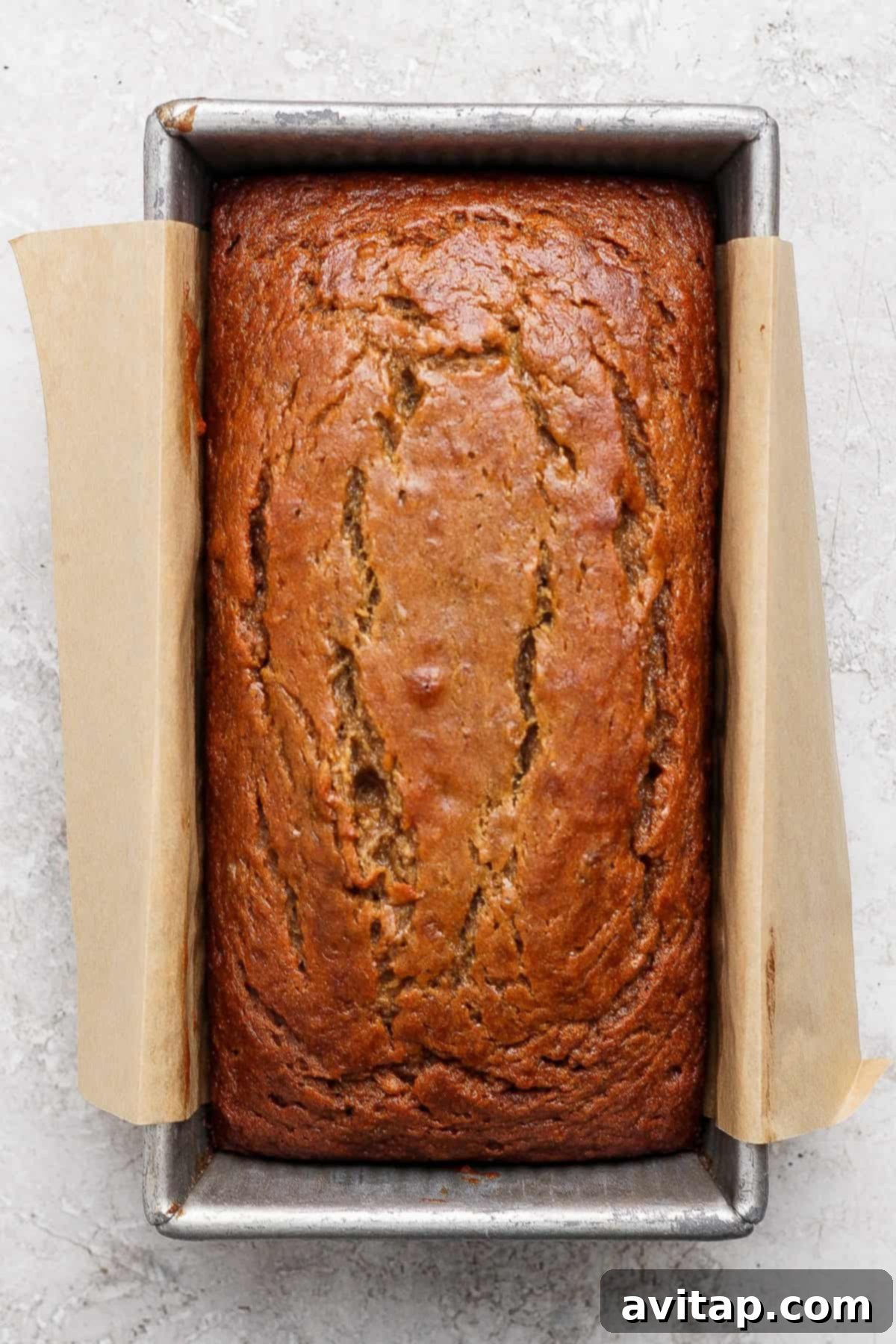 Chai-Spiced Brown Butter Banana Bread 5 A loaf of banana bread from a classic banana bread recipe sits in a rectangular metal pan lined with parchment paper, shown from above on a light textured surface.