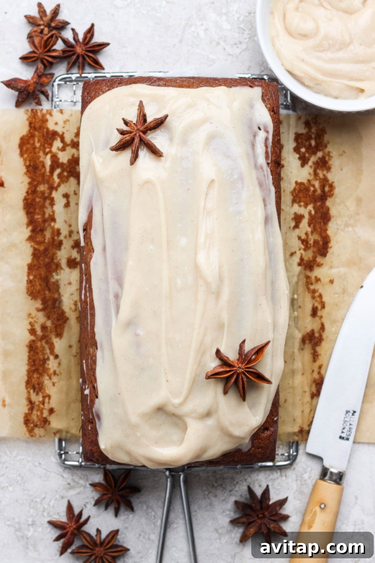 A delicious loaf cake of banana bread, featuring a delectable light brown frosting and elegantly garnished with whole star anise. It is presented on parchment paper with a knife and a small bowl of extra frosting nearby, ready to be enjoyed.