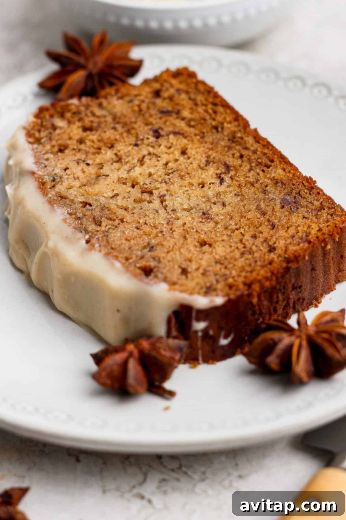Chai-Spiced Brown Butter Banana Bread 7 A slice of spiced banana bread loaf with white glaze on one edge, garnished with whole star anise, sits elegantly on a white plate—a delicious twist on the classic banana bread recipe.
