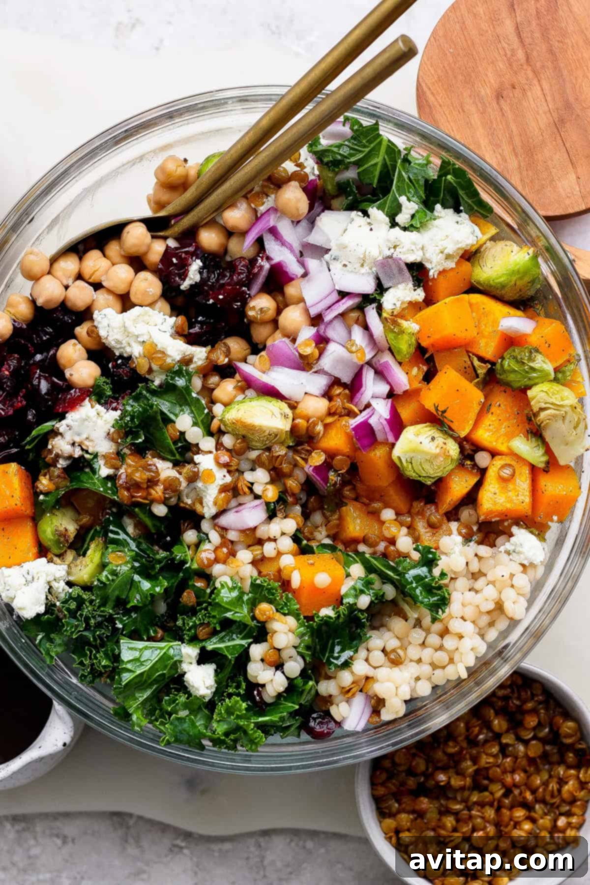 A vibrant couscous salad recipe featuring kale, chickpeas, roasted butternut squash, Brussels sprouts, red onion, blue cheese, dried cranberries, and lentils in a glass bowl with gold utensils on top.