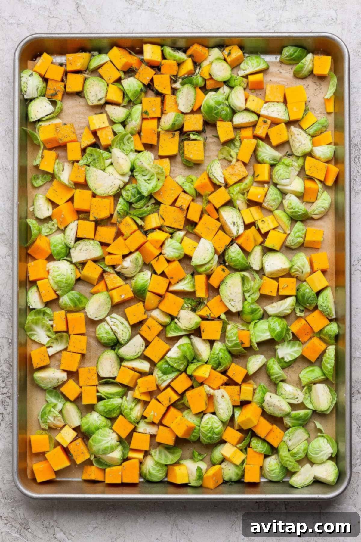 Cubed butternut squash and halved Brussels sprouts spread on a parchment-lined baking sheet, seasoned with salt, pepper, and herbs—perfect for adding to your favorite couscous salad recipe after roasting.