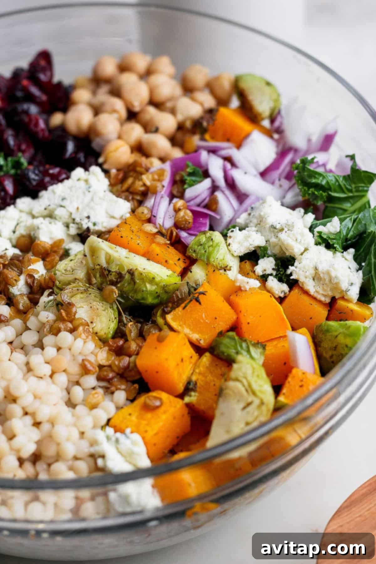 A glass bowl filled with a vibrant couscous salad recipe featuring chickpeas, dried cranberries, red onions, kale, roasted squash, Brussels sprouts, lentils, and crumbled cheese.