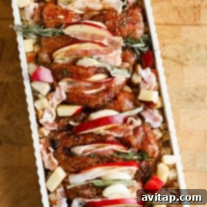 Raw seasoned chicken pieces topped with apple slices and herbs, arranged in a rectangular baking dish with cubed bread and chopped apples.