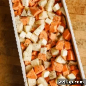A rectangular baking dish filled with cubed sweet potatoes and white potatoes, topped with small cubes of butter and sprinkled with seasoning, on a wooden surface.