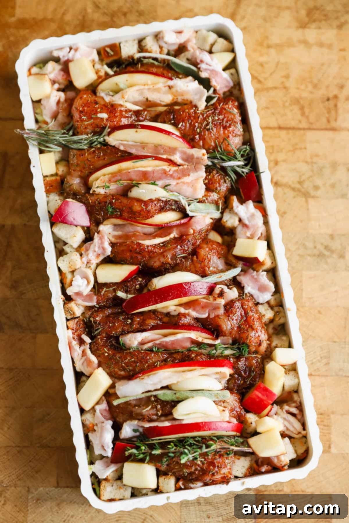 Raw seasoned chicken pieces topped with apple slices and herbs, arranged in a rectangular baking dish with cubed bread and chopped apples.