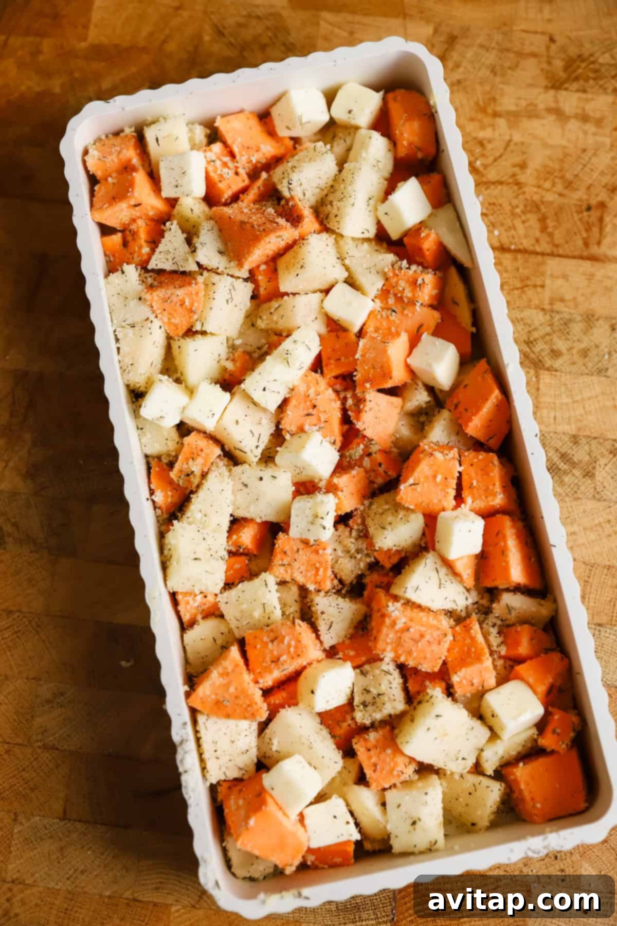 A rectangular baking dish filled with cubed sweet potatoes and white potatoes, topped with small cubes of butter and sprinkled with seasoning, on a wooden surface.