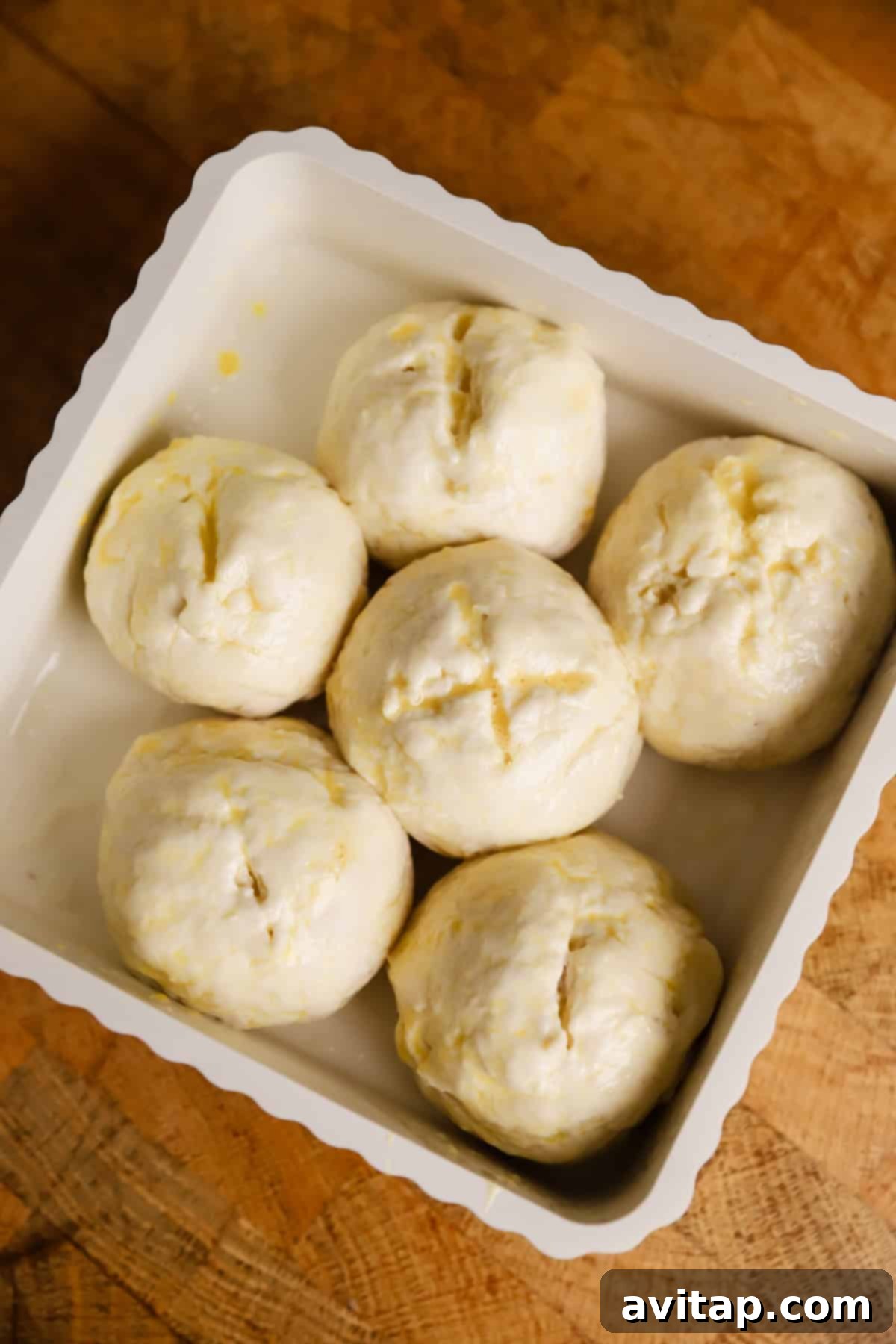 Six unbaked dough balls evenly arranged in a white baking dish, placed on a wooden surface.