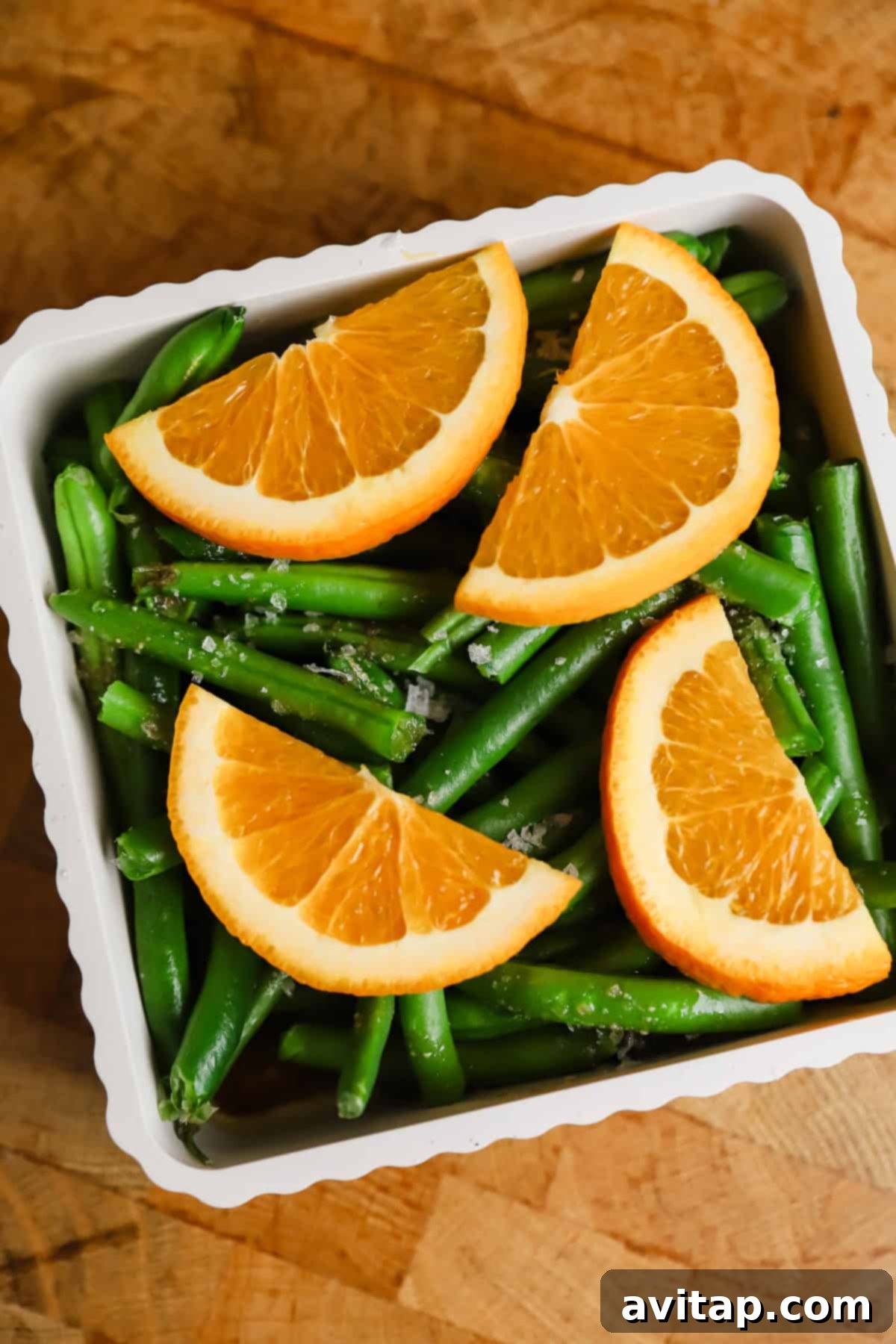 A white dish filled with green beans, topped with four orange slices, set on a wooden surface.