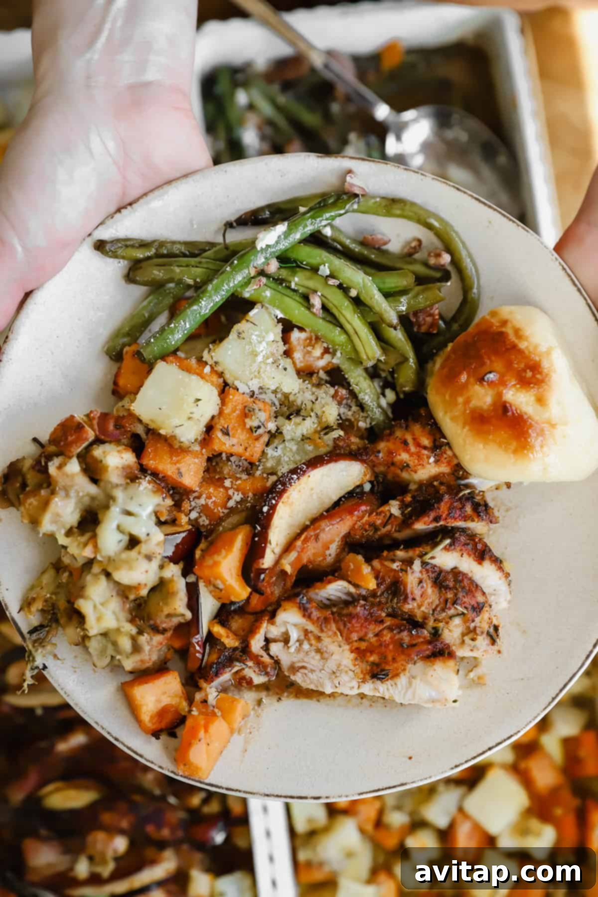 A plate with roasted turkey, green beans, stuffing, roasted vegetables, and a dinner roll, held over a table with more food trays.