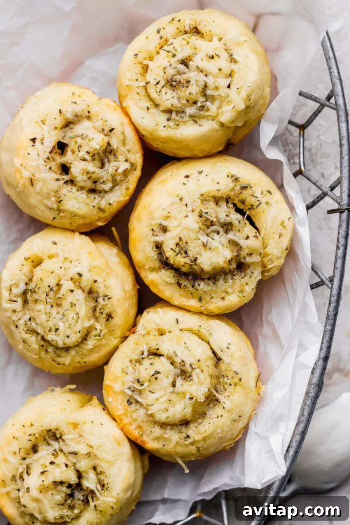 Six golden-brown, spiral-shaped bread rolls topped with herbs and shredded cheese, arranged on white parchment paper in a wire basket, ready to be served.