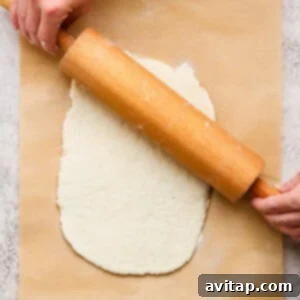 A person uses a wooden rolling pin to flatten dough on a sheet of parchment paper placed on a textured surface, demonstrating the rolling step.