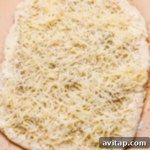 Unbaked flatbread dough topped with shredded cheese and herbs on a sheet of parchment paper, showing the seasoned dough before rolling.