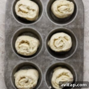 Six unbaked rolled dough pieces placed in a metal muffin tin, ready for baking, on a textured light surface, showcasing the rolls before entering the oven.