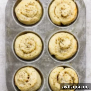 A metal muffin tray holding six unbaked, spiral-shaped dough rolls topped with herbs and shredded cheese on a light textured surface, prior to baking.