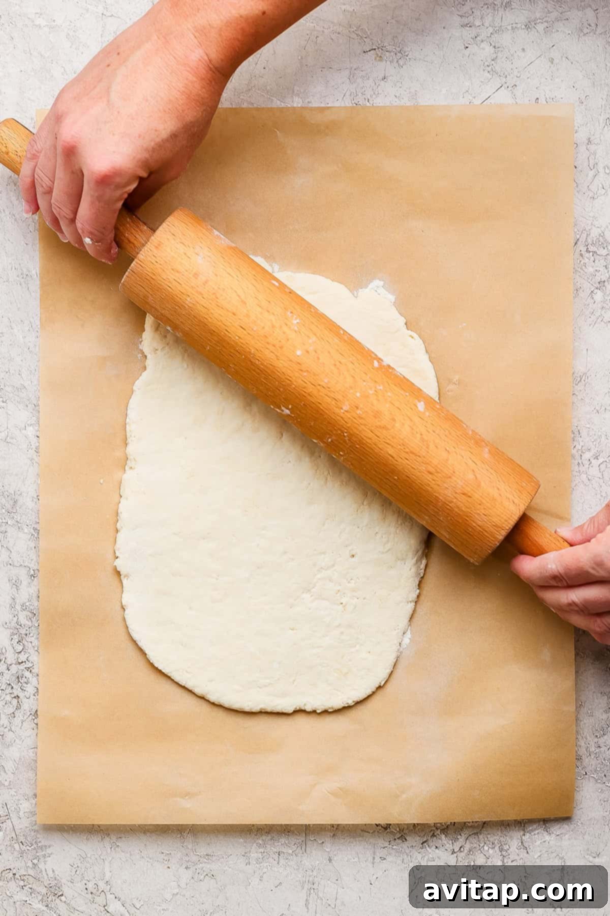 A person uses a wooden rolling pin to flatten dough on a sheet of parchment paper placed on a textured surface, preparing the base for the rolls.
