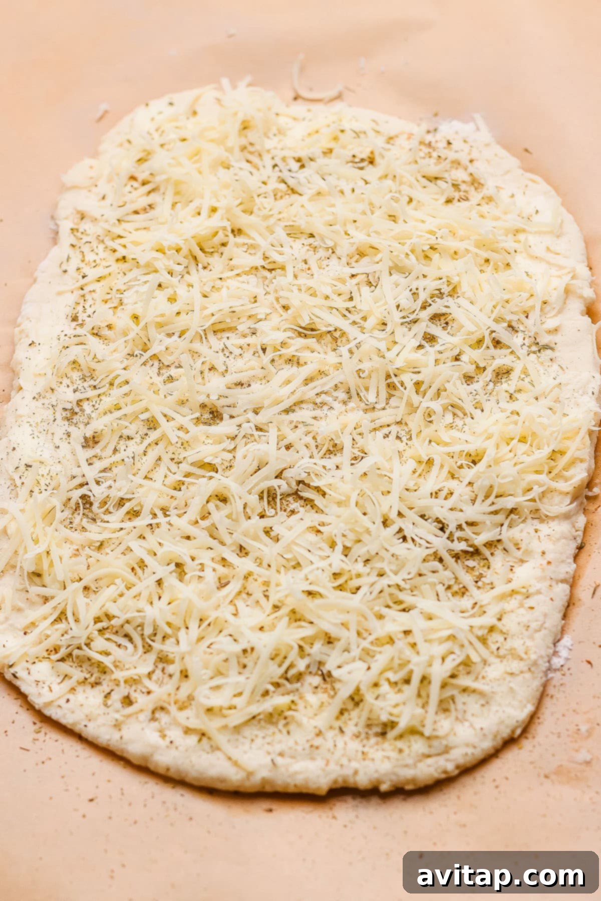 Unbaked flatbread dough generously topped with shredded cheese and a sprinkling of herbs on a sheet of parchment paper, ready to be rolled.