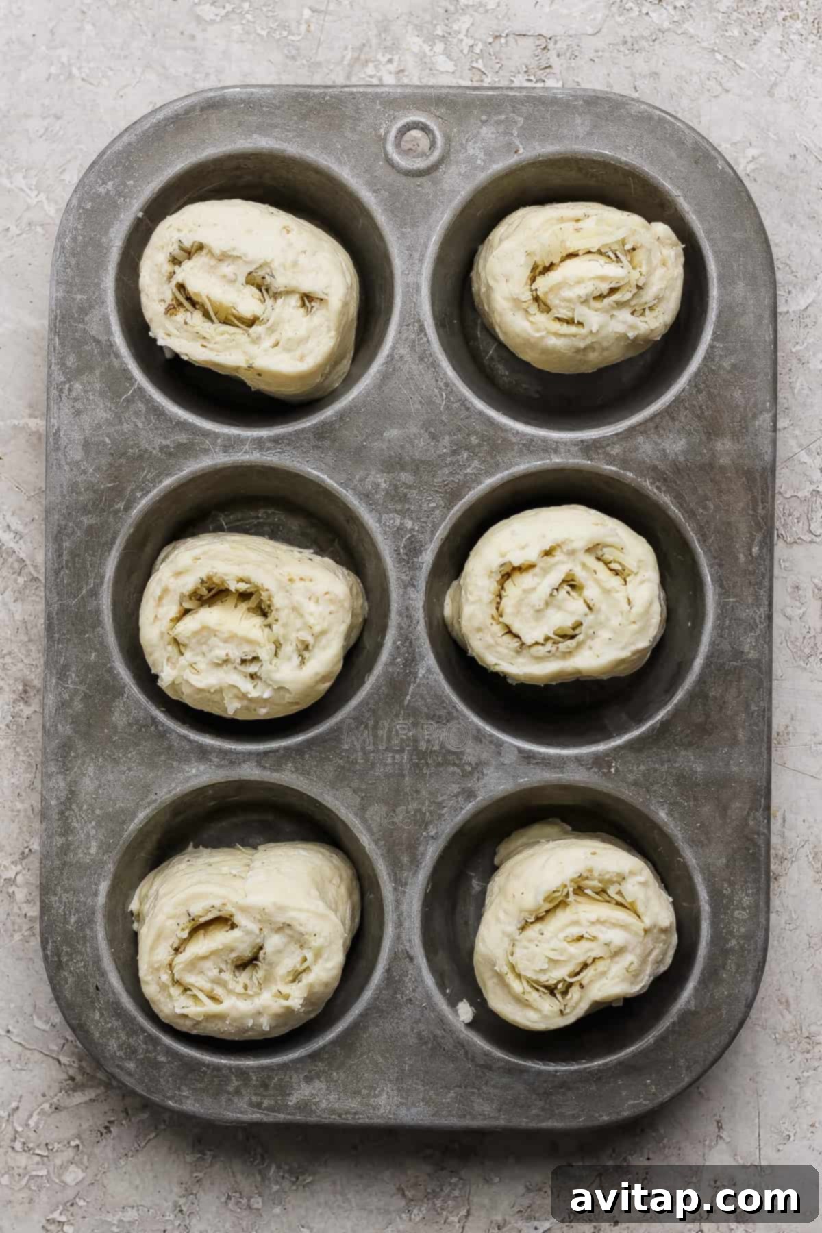Six unbaked rolled dough pieces, spiral-shaped and seasoned, neatly placed in a metal muffin tin, awaiting baking on a textured light surface.