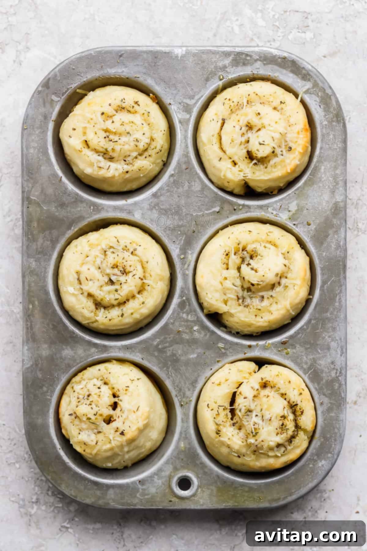 A metal muffin tray holding six unbaked, spiral-shaped dough rolls, glistening with butter and topped with herbs and shredded cheese, ready for the oven.